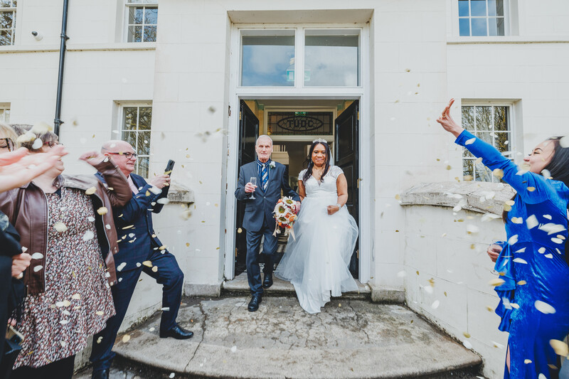 Newlyweds exit a white building doorway while guests throw confetti in celebration.