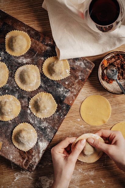 Hands crimping pastry edges in a muffin tin with flour-dusted surface and ingredients nearby.