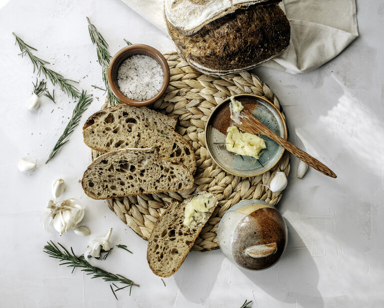 Flat lay of artisan bread, butter, salt, and fresh rosemary arranged on a natural surface.