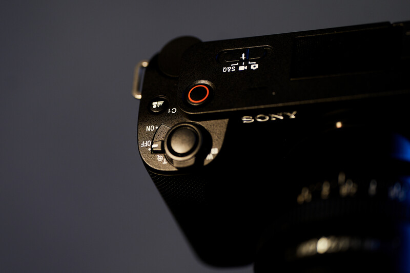 Close-up of Sony camera body showing control dial and red record button against gray background.