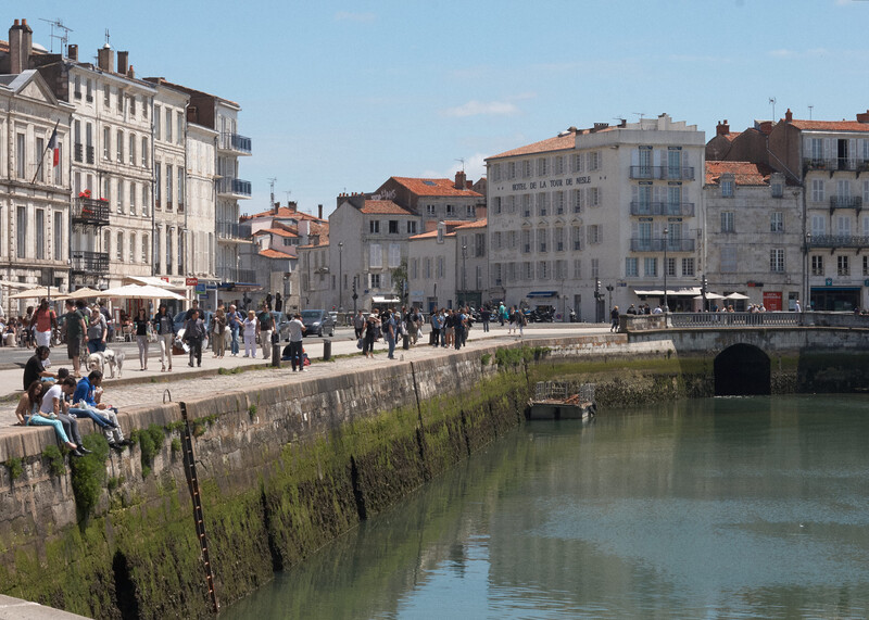 Waterfront plaza with stone quay, pedestrians, and European-style buildings reflected in calm water.