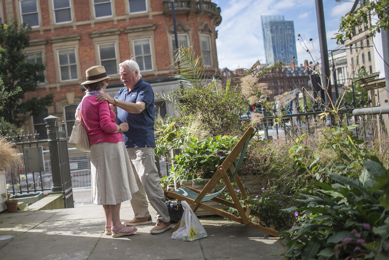 Two people conversing in a rooftop garden with urban architecture visible in the background.