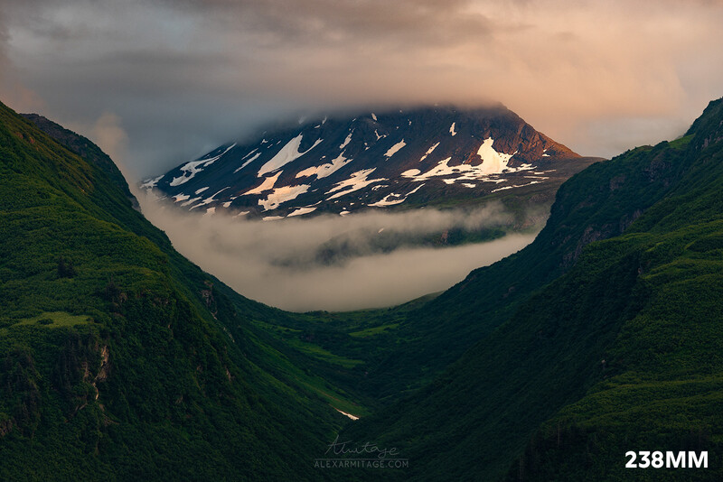 Snow-capped mountain framed by verdant valley walls beneath dramatic cloudy sky.
