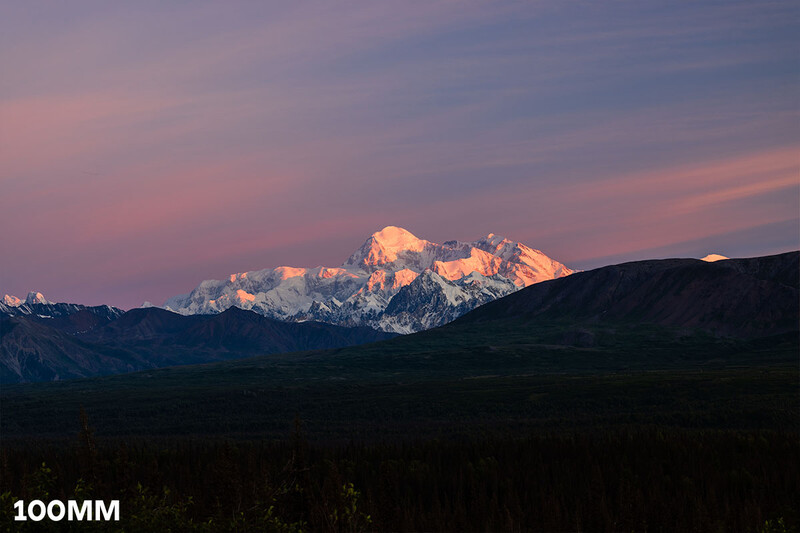 Snow-covered mountain peak glowing in alpenglow light against a purple twilight sky, with forested foothills silhouetted in the foreground.