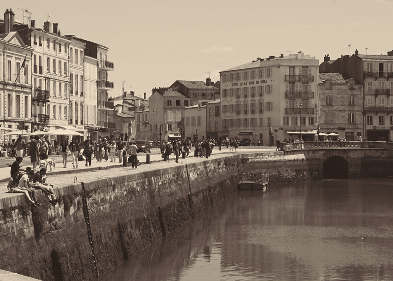 Historic European riverfront with stone embankment, period buildings, and pedestrians in 19th-century clothing.