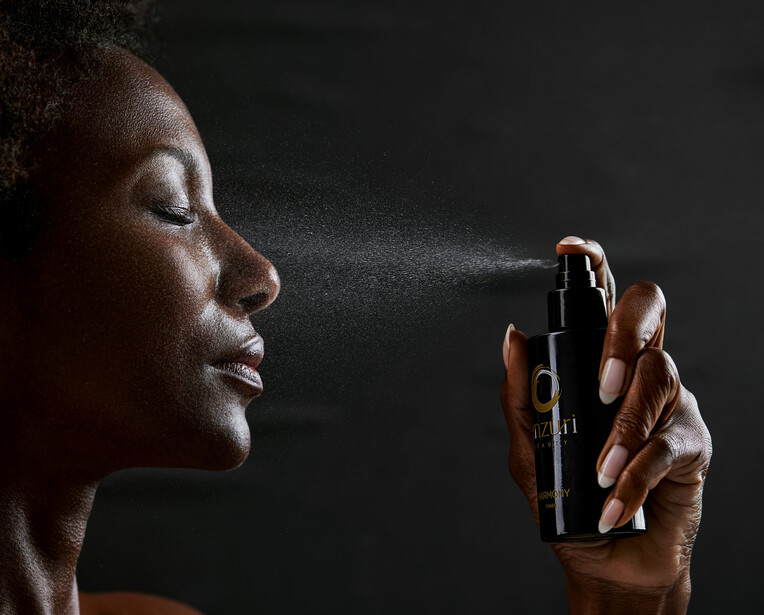 Woman in profile spraying facial mist or setting spray, with water droplets visible against dark background.
