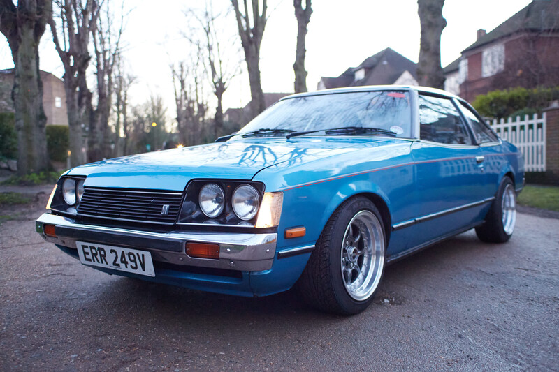 Bright blue coupe parked on a residential driveway with bare trees and houses visible in the background.