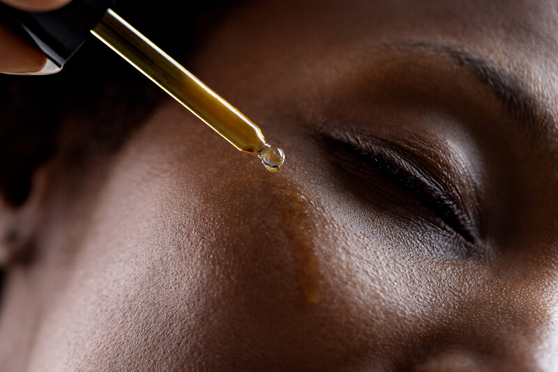 Dropper applicator dispensing golden liquid onto person's cheekbone with closed eye visible.