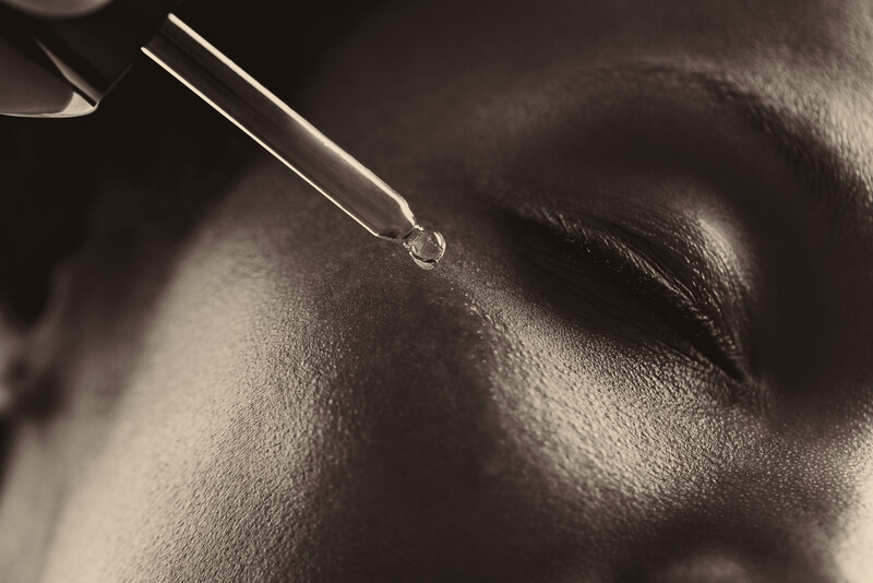 Dropper dispensing clear liquid onto textured skin with dramatic side lighting.