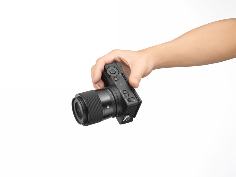 Hand holding a mirrorless camera with attached lens against white background.