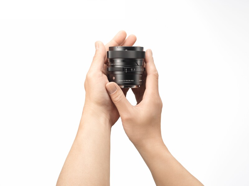 Hands holding a compact camera lens with focus ring and aperture markings visible against a white background.