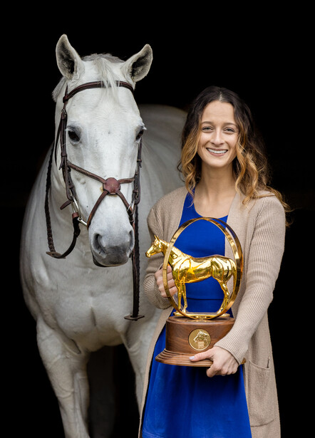 Woman in blue dress and cardigan holding a trophy next to a gray horse against a black background.