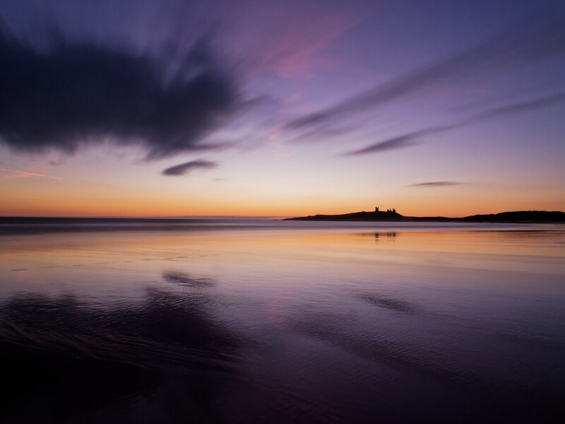 Calm bay at twilight with silhouetted islands against purple and gold sky.