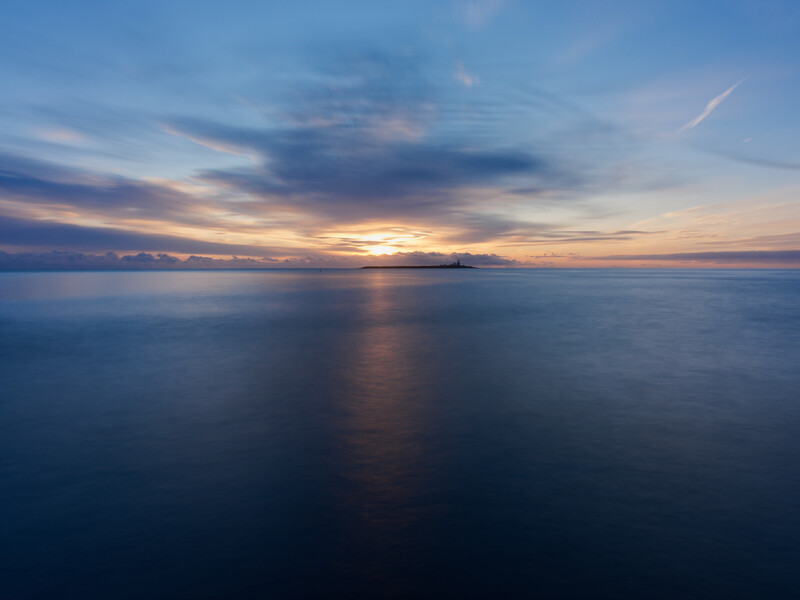 Serene seascape at sunset with golden light reflecting across calm water beneath a dramatic cloudy sky.
