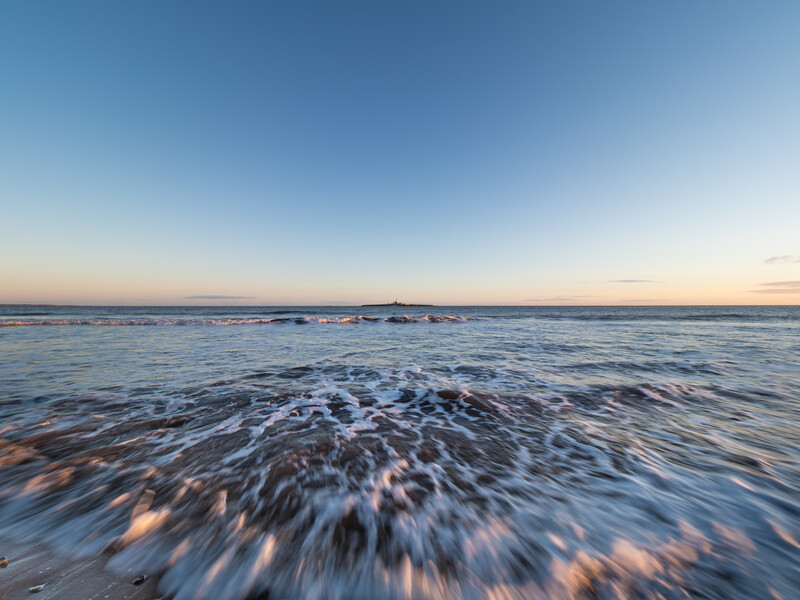Ocean waves washing onto a sandy beach at sunset with a clear blue sky and distant horizon.