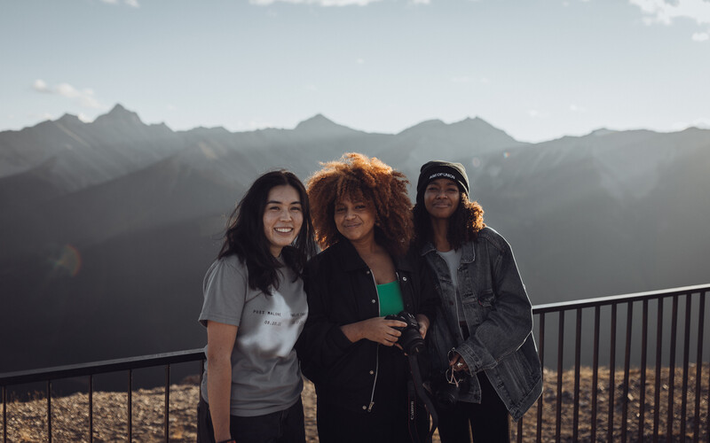 Three women posing together on a mountain overlook railing with dramatic peaks in the background.