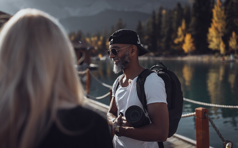 Man in white shirt and cap holding camera, conversing with woman with blonde hair by a lake at sunset.