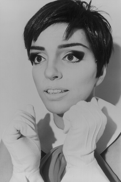 Black and white portrait of a person with short dark hair and defined eye makeup, chin resting on hands.
