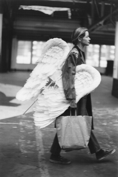 Woman holding large white feathered wings stands in an industrial space, photographed in black and white.