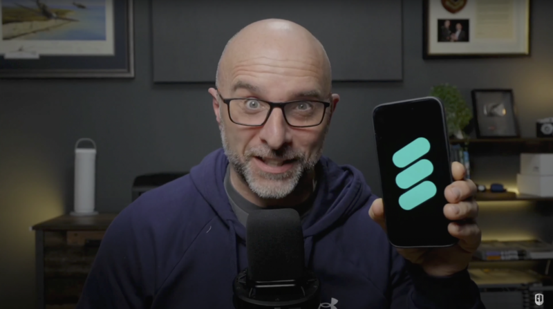 Man with glasses holding smartphone displaying app interface while speaking into microphone in home office setting.
