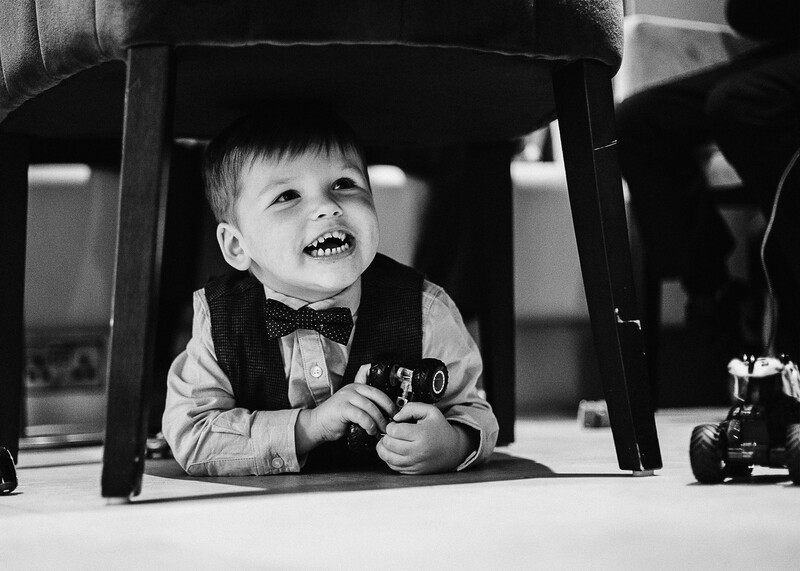 Young child in formal wear smiling beneath an overturned chair, holding a toy car.