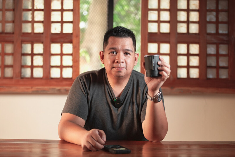 Man in gray v-neck shirt holding a compact camera while seated at a wooden table.