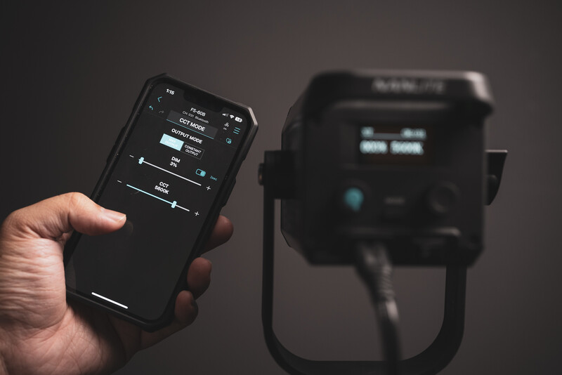Hand holding a wireless remote control with display screen, positioned in front of an out-of-focus studio light.