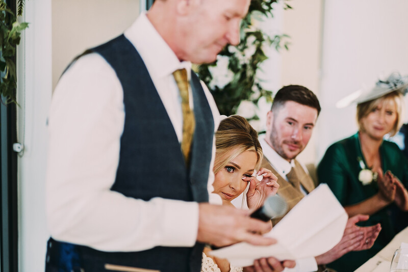 Wedding guests gathered indoors during a ceremony, with a man in vest and tie reading from a document while others look on.