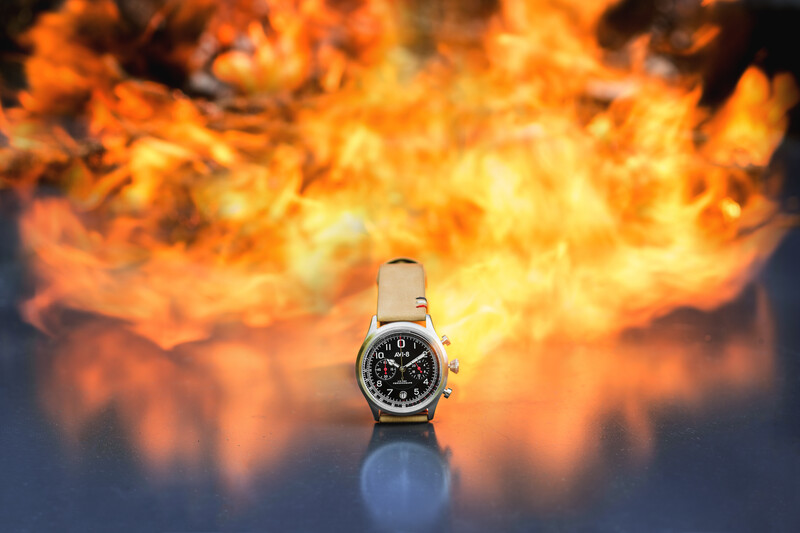 Analog chronograph watch with leather strap positioned against a blurred background of intense orange flames.