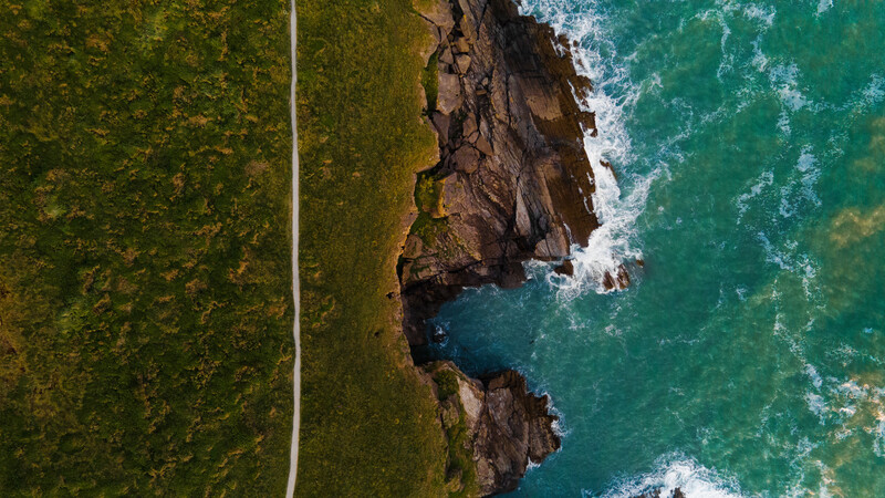 Aerial view of a dramatic rocky coastline with turquoise ocean waves and a straight road cutting through dense forest.