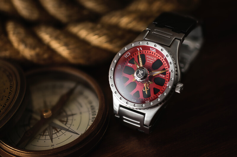 Wristwatch with red dial and metal bracelet beside a brass compass on dark fabric.