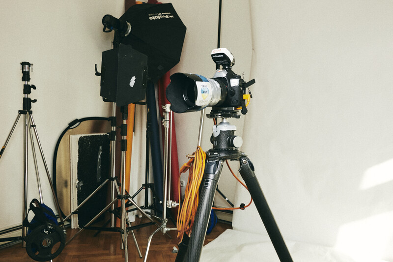 Photography studio setup with tripod-mounted telescope, lighting equipment, and black camera bag against white wall.