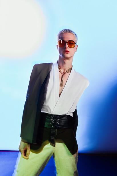 Man in blazer and neon green pants poses against blue gradient backdrop wearing sunglasses.