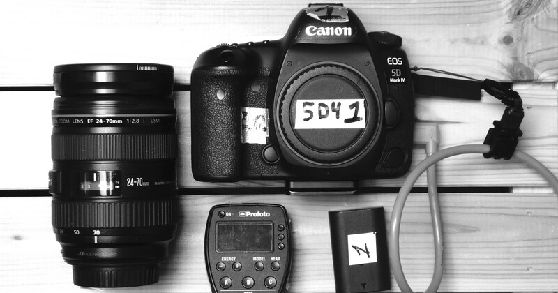 Canon EOS 5D Mark IV camera body with 24-70mm lens, Profoto flash trigger, and accessories arranged flat on white wood.