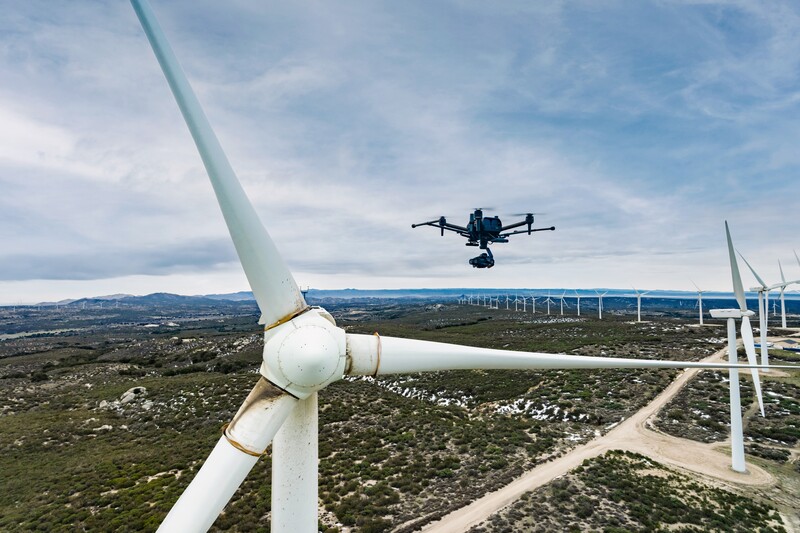 Drone hovering above white wind turbine in a landscape with multiple turbines visible in the distance.