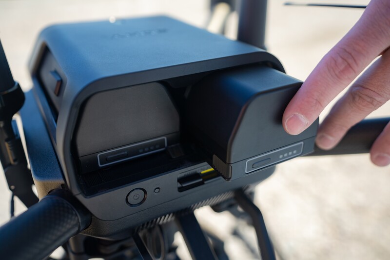 Hand opening the front panel of a blue and black drone to access the battery compartment.