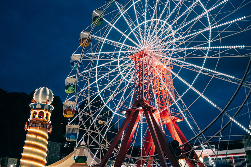 Illuminated Ferris wheel at night with glowing support structure and lit carnival rides alongside.