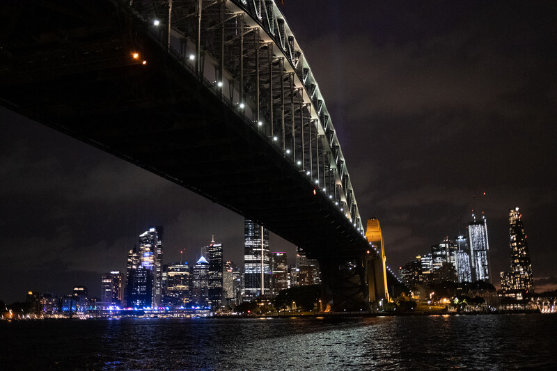 Sydney Harbour Bridge spanning dark water with illuminated city skyline behind it at night.