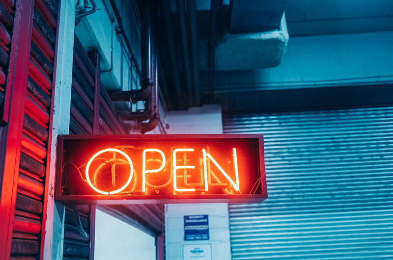 Bright red neon OPEN sign glowing against a corrugated metal storefront shutter and white wall.