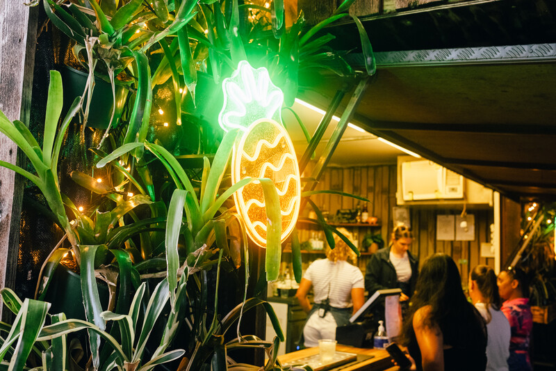 Neon pineapple sign glowing above storefront with tropical plants and customers visible inside at night.