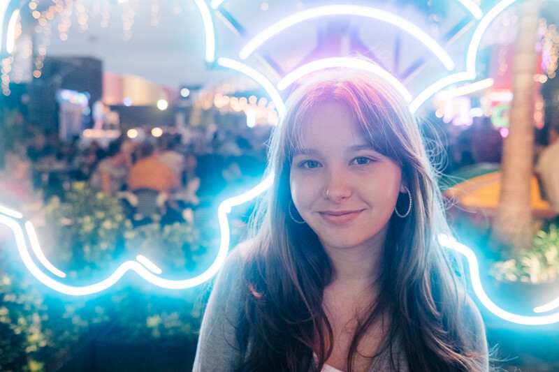 Young woman with long dark hair and hoop earrings smiling at camera, lit by glowing neon cloud-shaped signs.