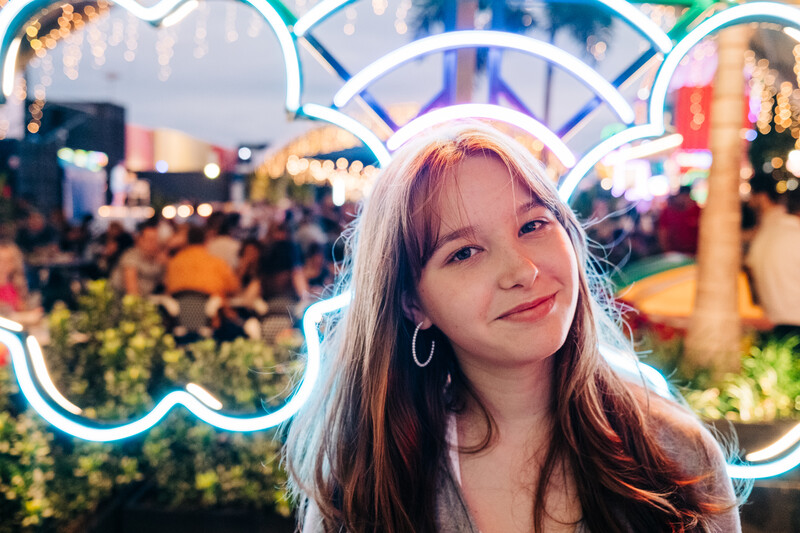 Young woman with long hair smiling at camera in front of glowing neon sign at night event.
