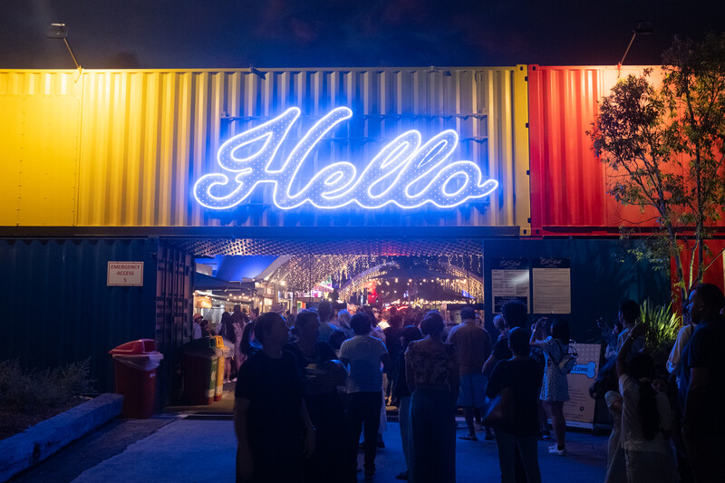 Neon blue "Hello" sign mounted on colorful corrugated metal shipping container storefront at night.