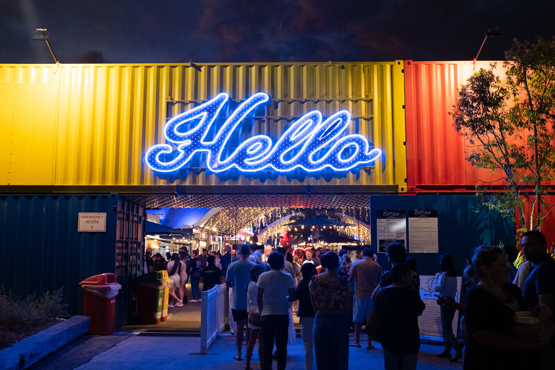 Neon blue "Hello" sign glowing above a crowded shipping container venue at dusk.