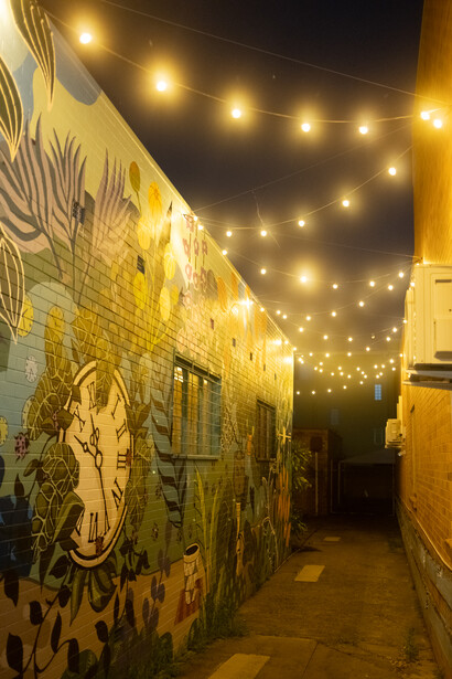 Narrow alleyway with yellow-painted walls decorated with black murals, illuminated by warm string lights overhead at night.