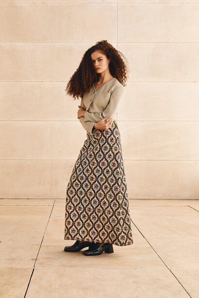 Woman in tan button-up shirt and patterned maxi skirt standing against beige tiled wall.