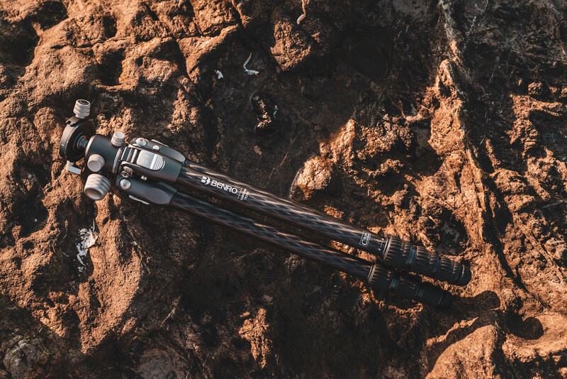 Tripod with telephoto lens positioned on rocky, textured ground terrain.