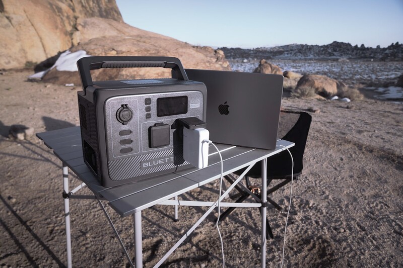 Portable power station and tablet displayed on camping table at beach location with rocky cliffs in background.