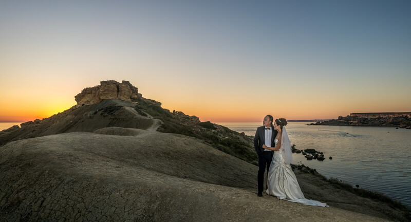 Bride and groom standing on a rocky outcrop overlooking a coastal bay at sunset.