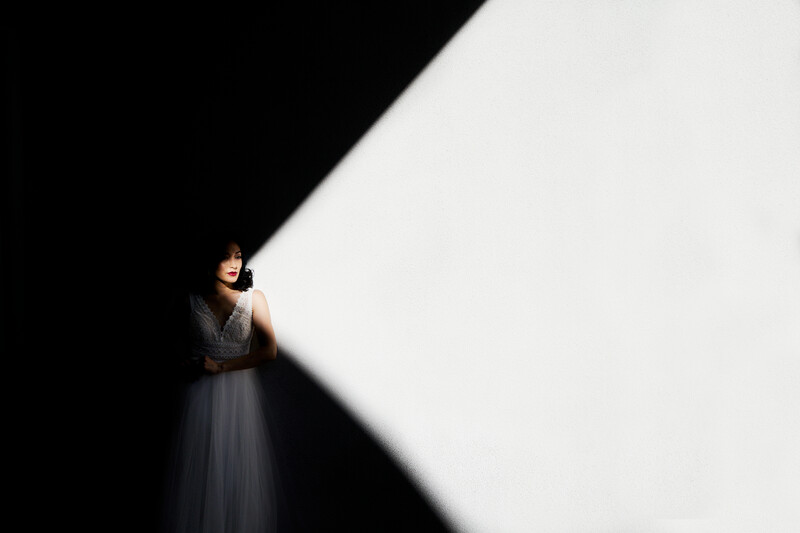 Woman in light-colored dress standing in dramatic shaft of light against dark background.